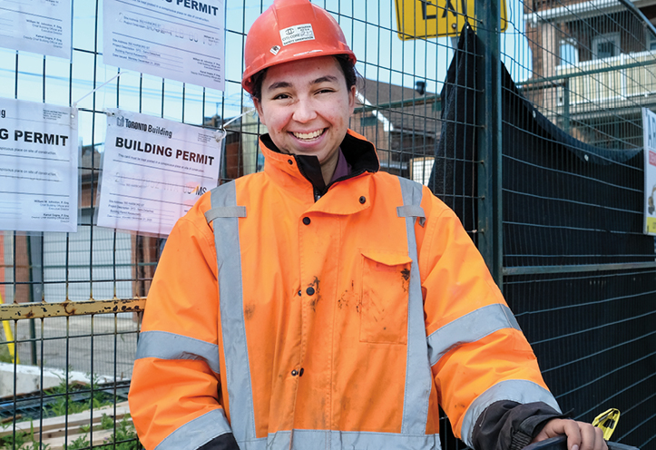 Woman smiling, wearing an orange construction vest and safety helmet